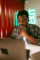 Focused young man working on laptop in modern workspace with neon exit sign during evening hours photo