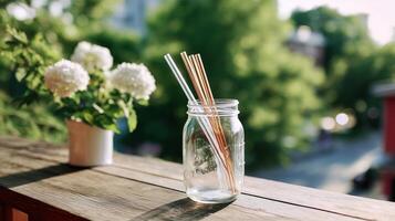 Glass jar holds reusable straws on wooden railing with potted flowers in a sunny outdoor setting photo