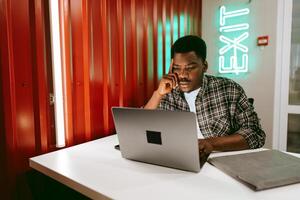 Focused individual working on a laptop at a modern workspace with neon exit sign in the background photo