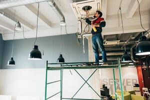 Worker performs maintenance on HVAC system while standing on scaffolding in an office space during the daytime photo