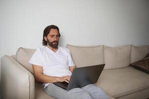Man Using Laptop While Sitting Comfortably at Home on a Beige Sofa photo