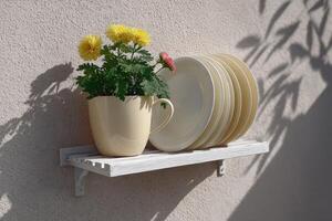 Cheerful flowers in a mug beside neatly arranged plates on a white shelf outdoors photo