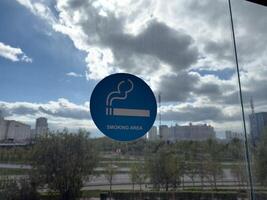 Blue smoking area sign on glass wall with urban view in background, illustrating the widespread normalization of smoking and growing demand for designated spaces in modern public environments photo