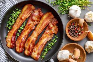 Fried bacon with thyme in pan and garlic with peppercorns on grey table photo