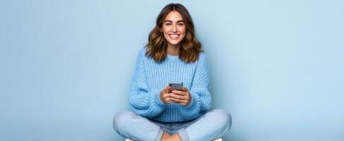 The smiling woman sitting casually while using her smartphone against a blue background. photo