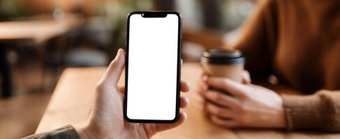 The smartphone held in a hand next to a coffee cup on a table. photo