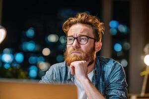 A bearded man stares thoughtfully at a laptop screen at nighttime photo