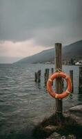 Lifeguard orange floatation device hanging on a wooden post at the water's edge on an overcast day photo