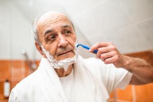 Handsome older man into the mirror while shaving his beard with razor, reflecting on a life of routine and care. photo