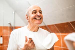 Content senior man with a towel in his hands checking and smiling at his reflection in mirror after washing his teeth and face in bathroom. photo