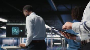 Close up of server room worker checking code on tablet next to colleague reviewing AI LLM visualization. Data center IT expert using device to monitor artificial intelligence systems, camera A video