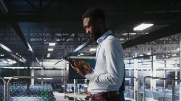 Technician walks in data center, doing maintenance tasks on tablet, ensuring optimal performance. IT expert reviewing diagnostics and applying patches to server hub gear, improving stability, camera A video