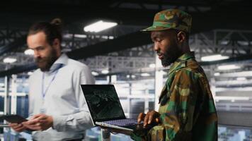 Military technician in data center monitoring warfare drone telemetry on laptop dashboard. IT specialist in army server room using tactical interface on notebook to support aerial operations, camera B video