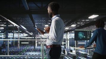 Admin on data center industrial platform analyzing infrastructure performance outputs on tablet. Server room employee doing job tasks, finetuning mainframes configurations, camera A video