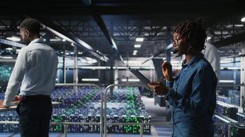 Server room admin using tablet and headset microphone to provide infrastructure tech support. Data center IT specialist resolves backend issues while coordinating with remote teams, camera A video