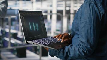 Close up of admin in data center using laptop to monitor neural network LLM visualization. Server hub IT expert overseeing rigs powering machine learning processes using notebook, camera B video