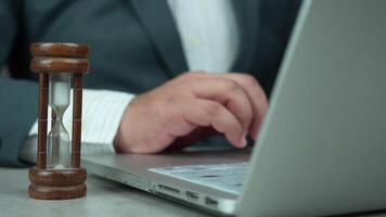 Male in suit hands are typing on the keyboard on a laptop, time is running on an hourglass nearby video