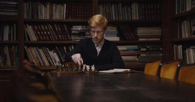A young man of European appearance, dressed in a business suit, is studying with concentration to play chess alone, using a textbook, sitting at a table photo