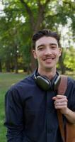 A young man with a backpack on his shoulders and headphones around his neck smiles while looking at the camera standing in a city park on a summer day photo