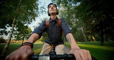 A young man with headphones and a backpack on his shoulders rides a bicycle in a city park. Front view. photo
