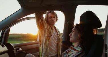 A woman is sitting in a car, a man is standing near the open door, they are smiling and talking. A sunset date in a field photo