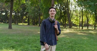 A young man with a backpack on his shoulders and headphones around his neck smiles while looking at the camera standing in a city park photo