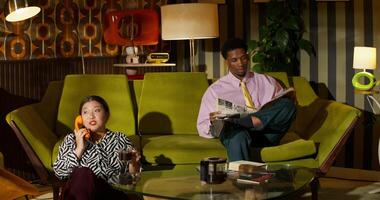 A woman smiles while talking on a wired phone. She is sitting on the floor next to a sofa where a man is reading photo