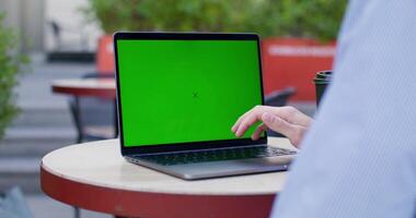 A man typing on a laptop with green screen sitting at a table on a cafe terrace. Close-up of his hands, an unrecognizable person. Copy space photo