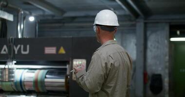 A man in a protective helmet and glasses uses a tablet while working at a factory. He stands near machine photo