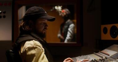 A sound engineer uses an audio mixer while recording a song in the studio. The rapper sings in the room behind the glass photo