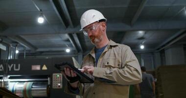 A man in a protective helmet and glasses uses a tablet while working at a factory. He stands near production machine photo