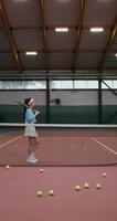 A young woman of Asian appearance in a short skirt comes to the tennis court with a tennis racket in her hands and leaning against a stretched tennis net posing looking at the camera, vertical photo