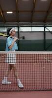 A young woman of Asian appearance in a short skirt comes to the tennis court with a tennis racket in her hands and leaning against a stretched tennis net posing looking at the camera photo