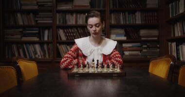 A young woman plays a chess game alone, thinking long concentratedly between moves, sitting at a table in the library photo