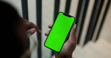 A mobile with a green screen in the hand of a man of African American appearance scrolling the screen, close-up, filming from behind. Copy space, mockup photo
