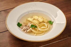 Chicken consomme soup with egg noodles and parsley in white bowl on rustic wooden table photo