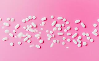 Many white pills are randomly arranged on a pink background in this top view photo