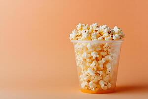 Fresh popcorn overflows from clear plastic cup against a solid peach colored background photo