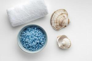 Bowl of blue bath salt sits beside towel and decorative seashells on white surface photo