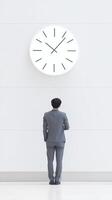 Businessman in gray suit standing in front of large wall clock, contemplating time and productivity in a modern office environment with minimalistic design photo