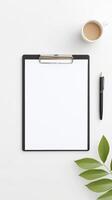 Clipboard with blank paper, black pen, green leaves, and coffee cup on a clean white surface, ideal for mock up and copy space design projects photo