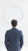 Man in formal suit standing with back to camera, gazing at large wall clock in minimalistic office space, reflecting on time and productivity photo