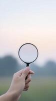 Hand holding a magnifying glass against a soft pastel sky, with blurred greenery in the background, symbolizing exploration, curiosity, and discovery in nature photo