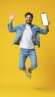 Happy man jumping and showing blank screen smartphone on yellow background photo