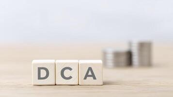 Wooden blocks displaying letters DCA are placed on a wooden surface, with stacks of coins in the background, symbolizing investment strategies and financial planning concepts photo