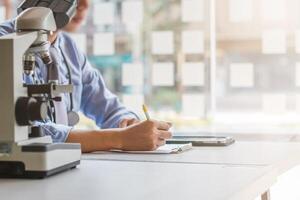 A man is writing on a piece of paper while sitting in front of a microscope photo