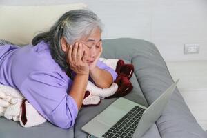Exhausted Senior Woman Resting While Using Laptop, Mature Woman with Fatigue Lying on Bed with Computer, Tired Elderly Lady Propping Her Head on Hands Looking at Screen photo