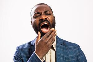 Portrait of a tired African American businessman in a suit yawning, with eyes closed, showing signs of exhaustion after a long day at work. Studio shot on gray background. photo