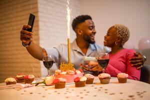 Smiling African boyfriend and girlfriend bonding during birthday celebration, surrounded by cake, wine and fireworks, sharing a joyful selfie at their home. Selective focus on cake. photo