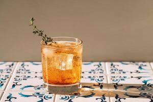 Close-up of a glass of soda and ice cubes with a sprig of thyme. Cool and delicious mineral water with fresh leaves. A sweet carbonated drink on a table with patterned tiles. photo